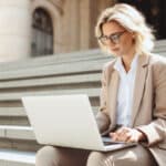 Woman sits on steps outside with her laptop to review board governance training.