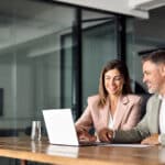 Two board members use a laptop equipped with hybrid board meeting tools to log in to a board meeting.